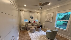 Ein Wohnzimmer mit Sofa, Hocker und Sessel, einem Deckenventilator und einem Fenster mit Blick auf Grünflächen.