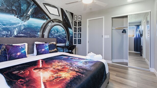 Une chambre à coucher avec un grand lit agrémenté d’une couvre-lit sur le thème de l’espace, un ventilateur de plafond et une vue sur un couloir avec un miroir et une porte.