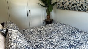 Et soveværelse med et mønstret sengetæppe, en potteplante, et skab og et blomsterbillede på væggen.