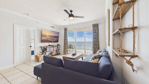 A living room with a sofa, a television, a bookshelf, and a view of the ocean through a sliding glass door. - Sunset Tides 1208-Gulf Front Balcony-Sleeps 6 (Panama City Beach)