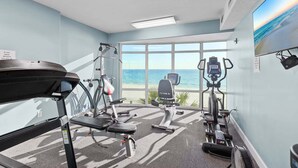 Une salle de sport équipée d’un tapis de course, d’un vélo elliptique et d’un vélo d’appartement, de grandes fenêtres offrant une vue sur l’océan et d’un ventilateur de plafond.