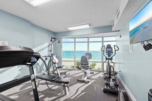 A gym with a treadmill, elliptical, and stationary bike, large windows with a view of the ocean, and a ceiling fan. - Sunset Tides 1208-Gulf Front Balcony-Sleeps 6 (Panama City Beach)