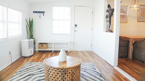 Une pièce avec un plancher en bois, une table ronde surmontée d’un vase, un banc blanc, un porte-manteau fixé au mur et une porte.