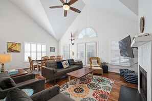 A spacious living room with a high ceiling, a ceiling fan, a fireplace, and a large window. - 418 - Nags Head Sound-side retreat with hot tub! (Nags Head)