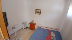 A bedroom with a bed, a radiator, a bedside table with a lamp, a picture on the wall, and a standing fan.