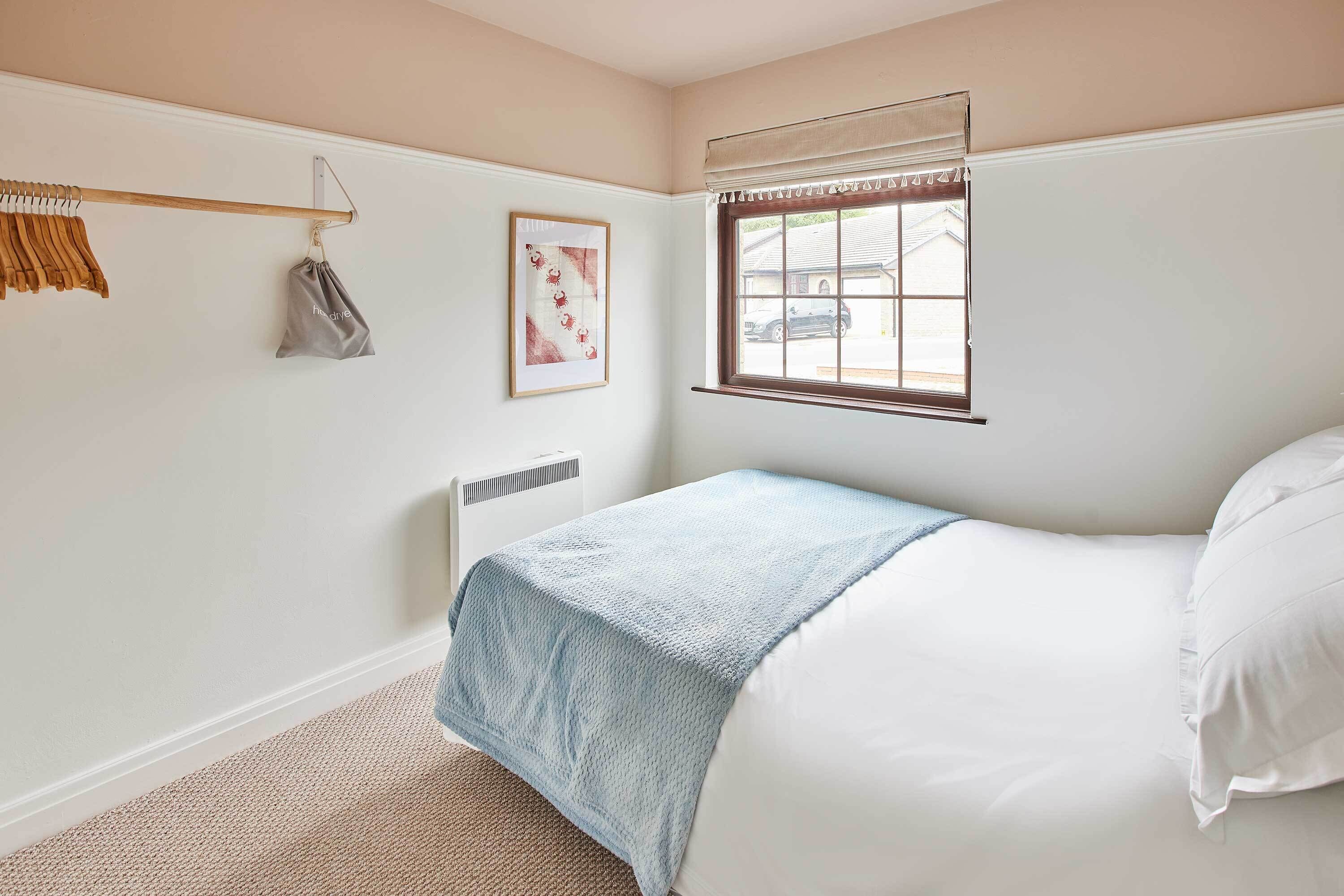 Un dormitorio con cama, ventana, radiador, perchero y un cuadro en la pared.