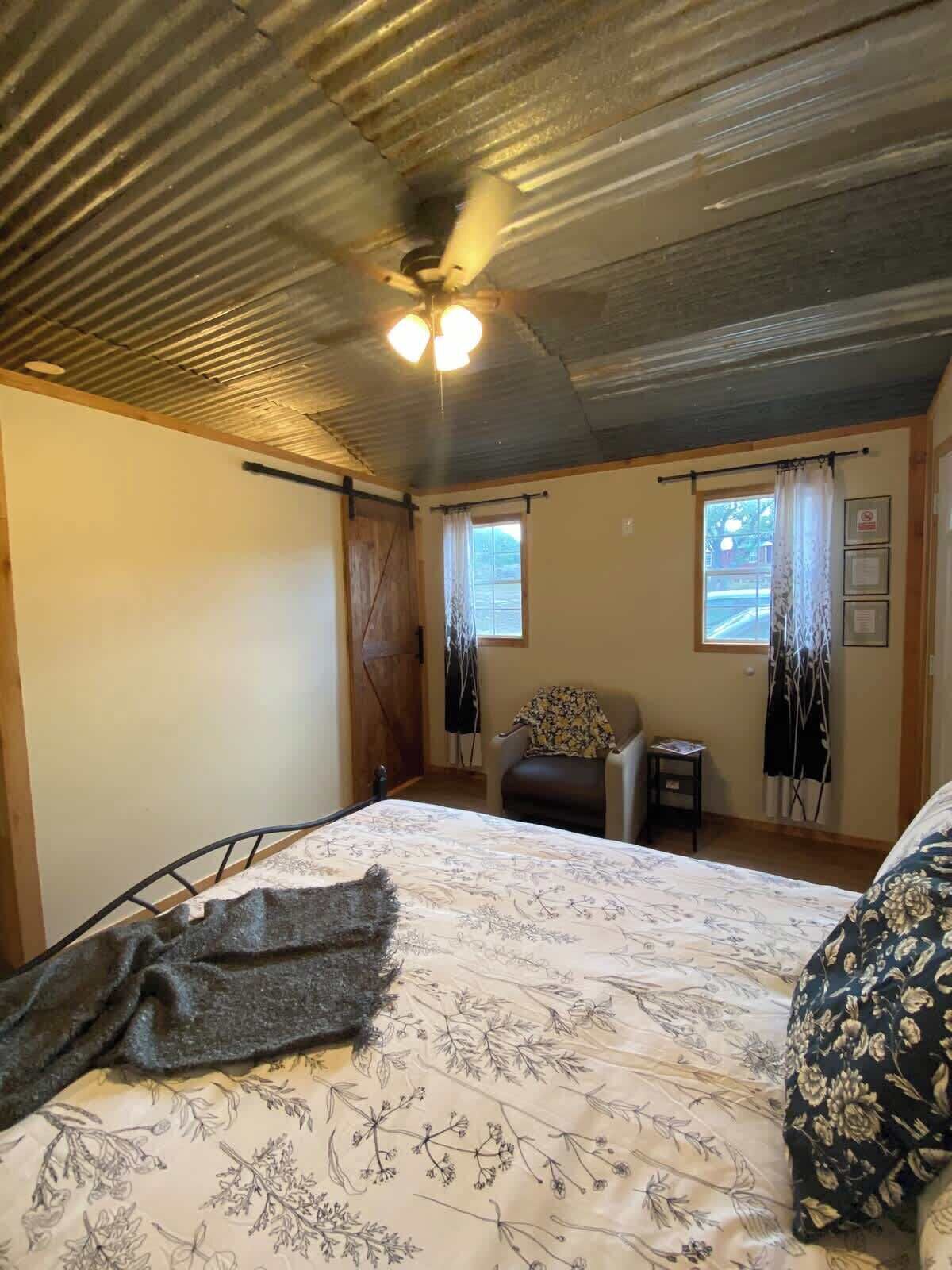 Un dormitorio con cama, una silla, una mesita y un ventilador de techo.