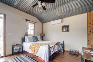 Een slaapkamer met een bed, nachtkastje, lamp, raam met gordijnen, plafondventilator en een airconditioner.
