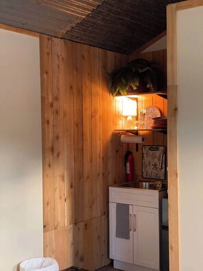 A small, compact kitchen with wooden walls, a white cabinet, and a shelf with decorative items. - Hill Country Cabin 2 (Canyon Lake)