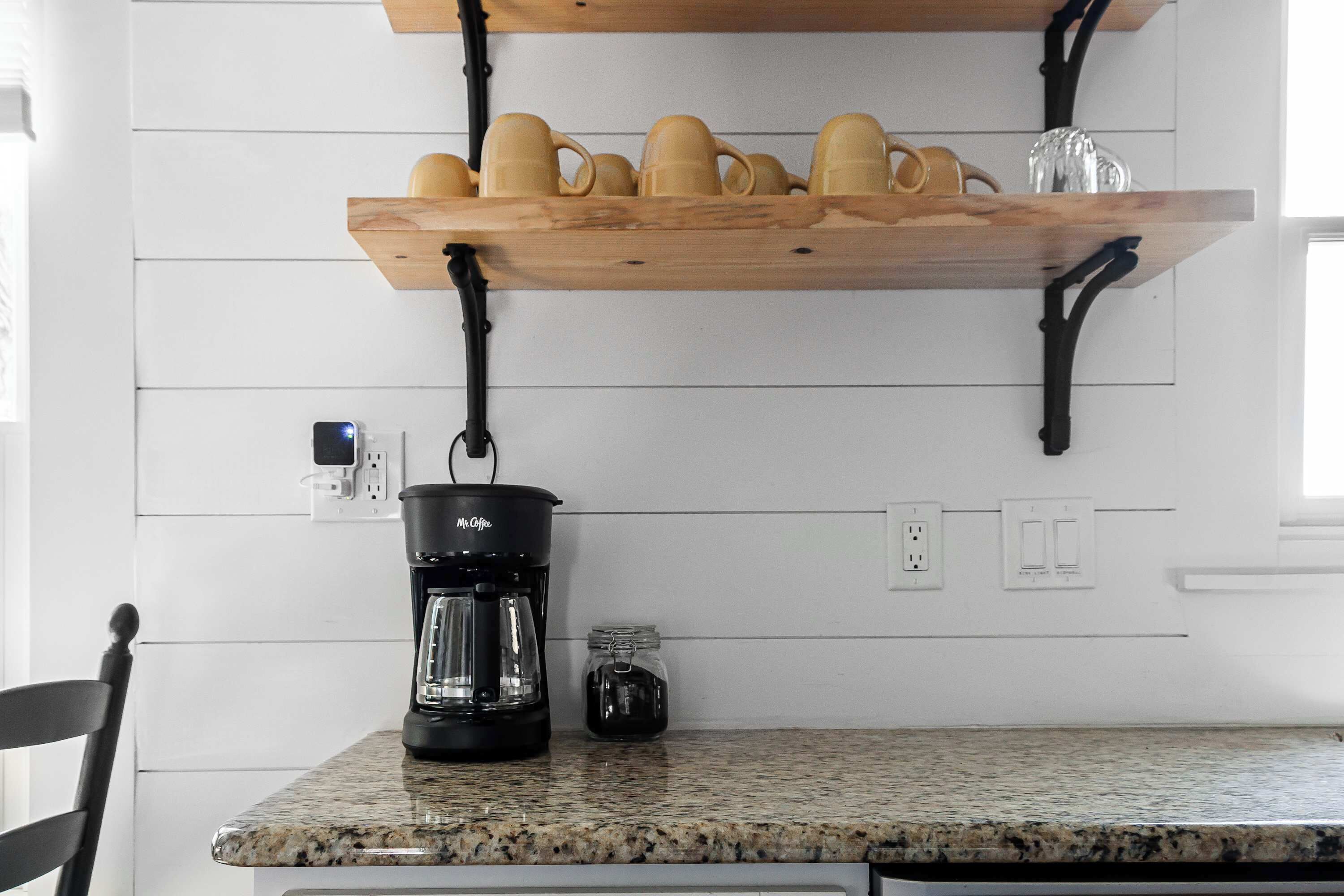 Una cocina con encimera de granito, cafetera, tarro y estante con tazas.