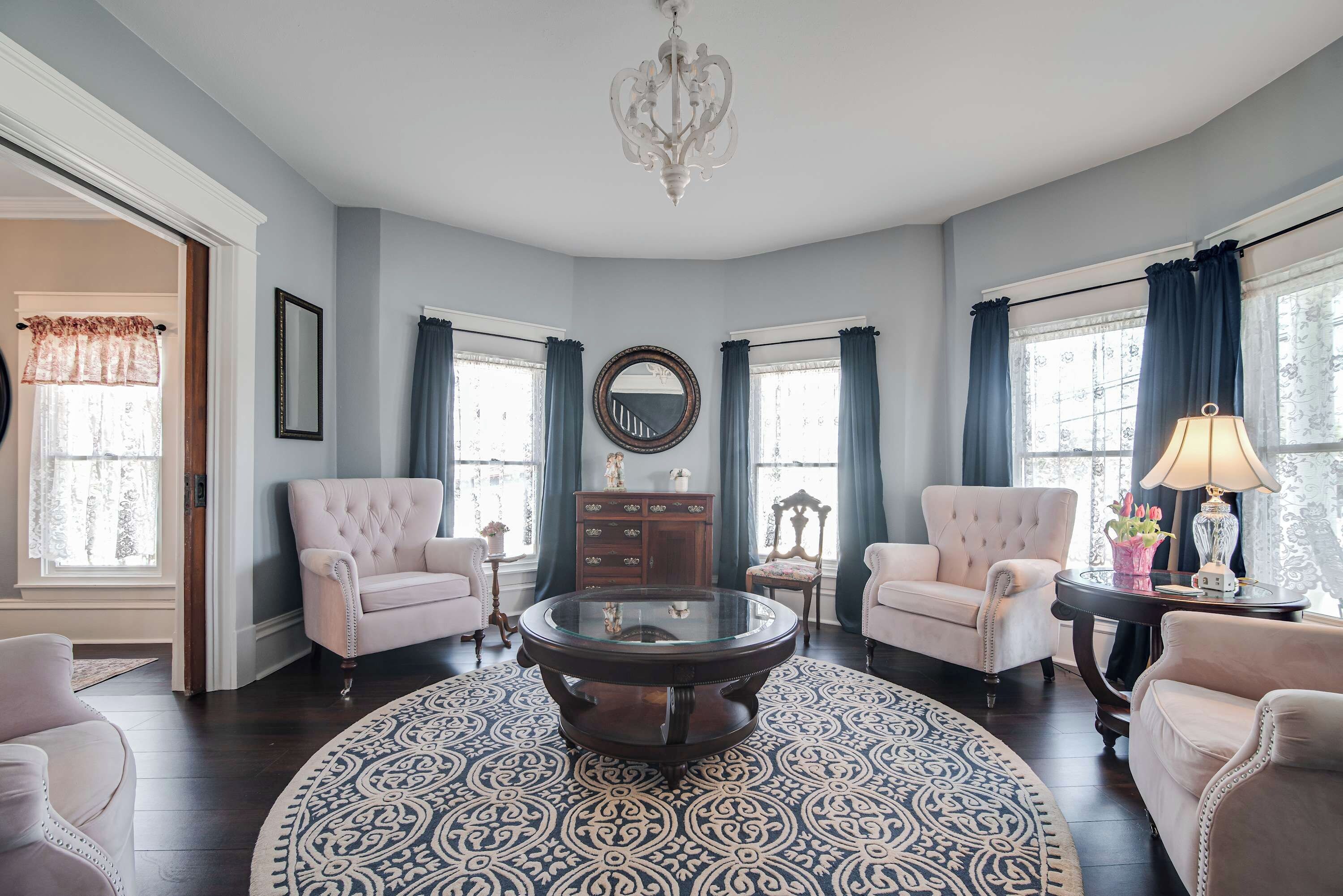 A spacious living room with a round glass coffee table, two armchairs, a wooden cabinet, and a chandelier.
