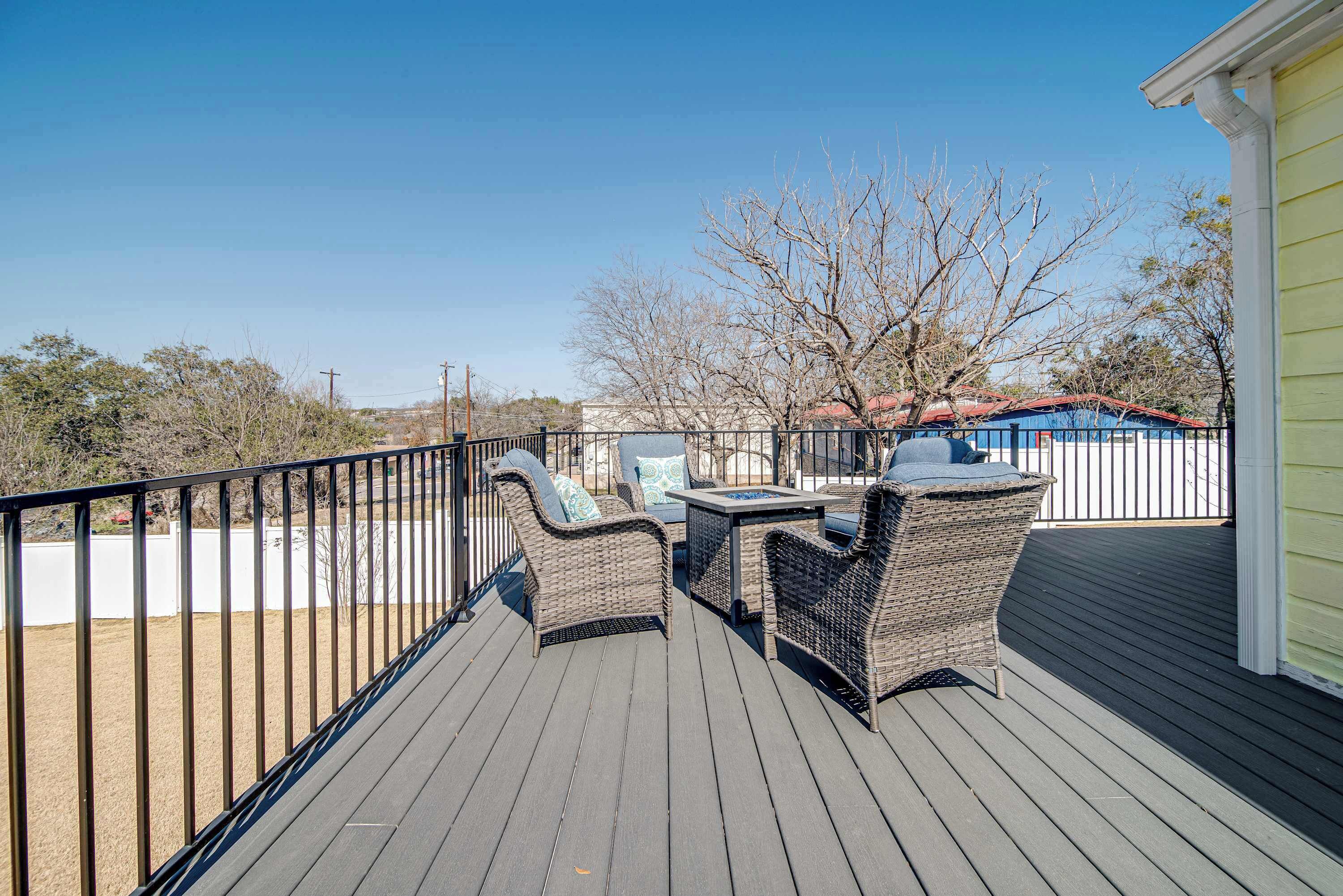 Una terraza de madera con muebles de mimbre y una mesa, con vista a un patio cercado y un edificio con techo rojo.