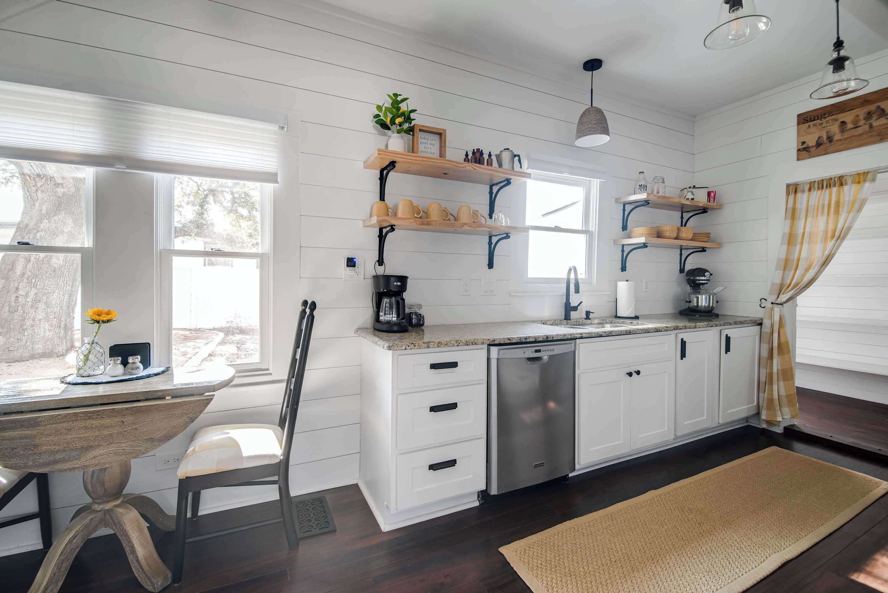 A kitchen with white cabinetry, a dishwasher, and a countertop with various kitchen items. There is a window with blinds, a chair, and a rug on the floor.