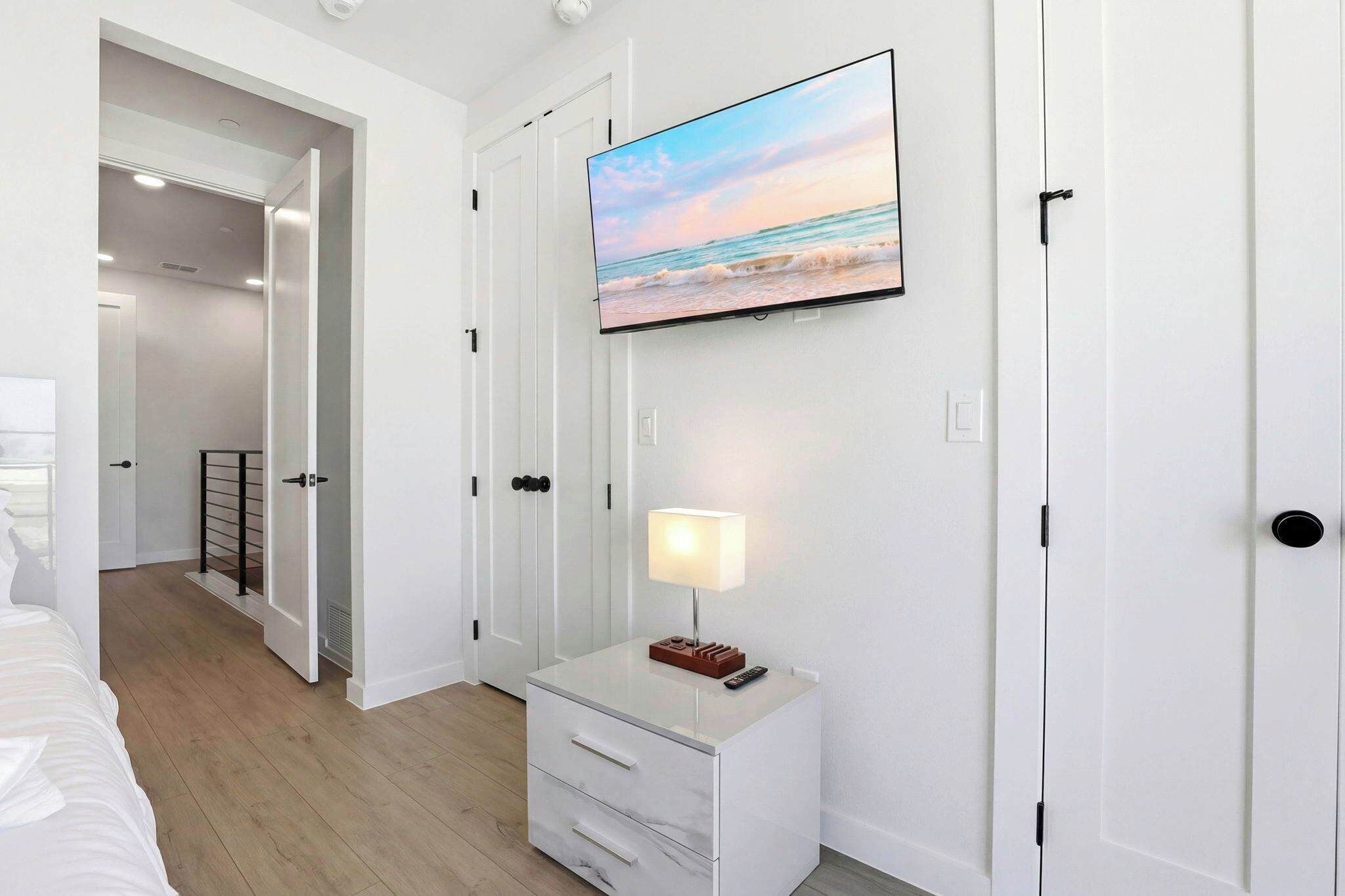 Un dormitorio con cama, una mesita de noche con lámpara, un televisor en la pared y una puerta que da a otra habitación.