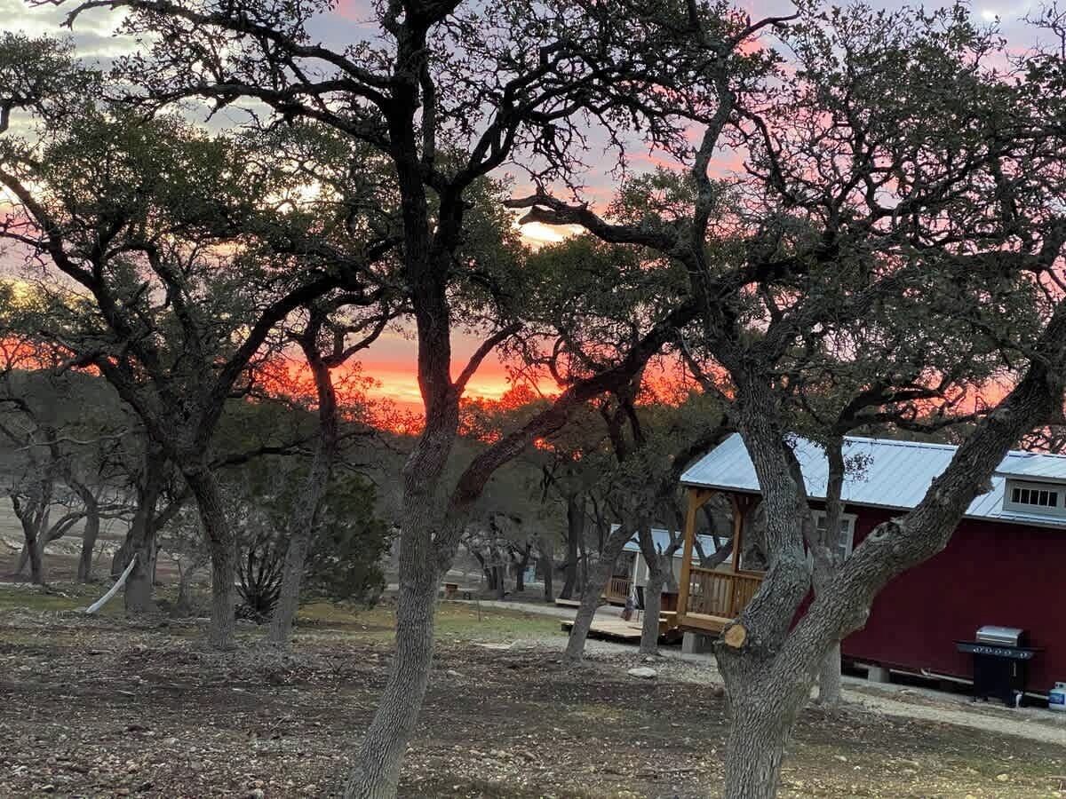 Een rustieke hut met een houten terras en rode gevel, omringd door bomen en een zonsondergang op de achtergrond.