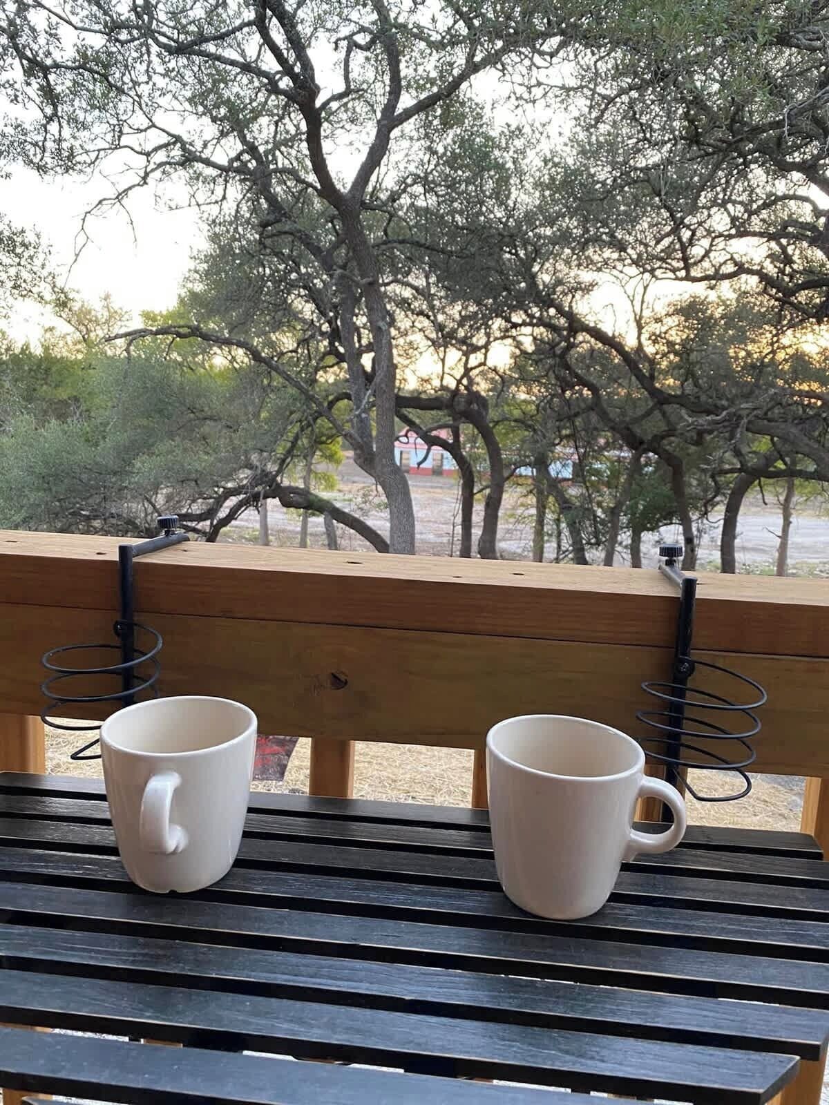 A wooden bench with two white mugs on it, overlooking a natural landscape with trees and a distant building.