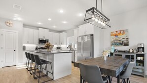 A modern kitchen with white cabinets, a central island with bar stools, a stainless steel refrigerator, and a hanging light fixture. - Southern Hillcountry Hideaway (Canyon Lake)