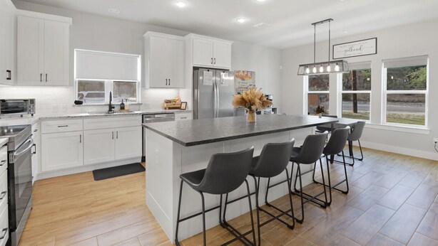 A modern kitchen with white cabinets, a central island with bar stools, a stainless steel refrigerator, and a hanging light fixture. - Southern Hillcountry Hideaway (Canyon Lake)
