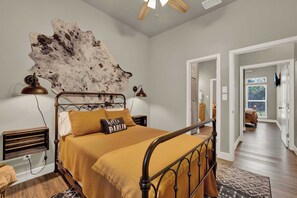 Een slaapkamer met een metalen bedframe, een grote wandlamp, een houten hoofdbord, een bed met een mosterdkleurige deken en een wandheater.