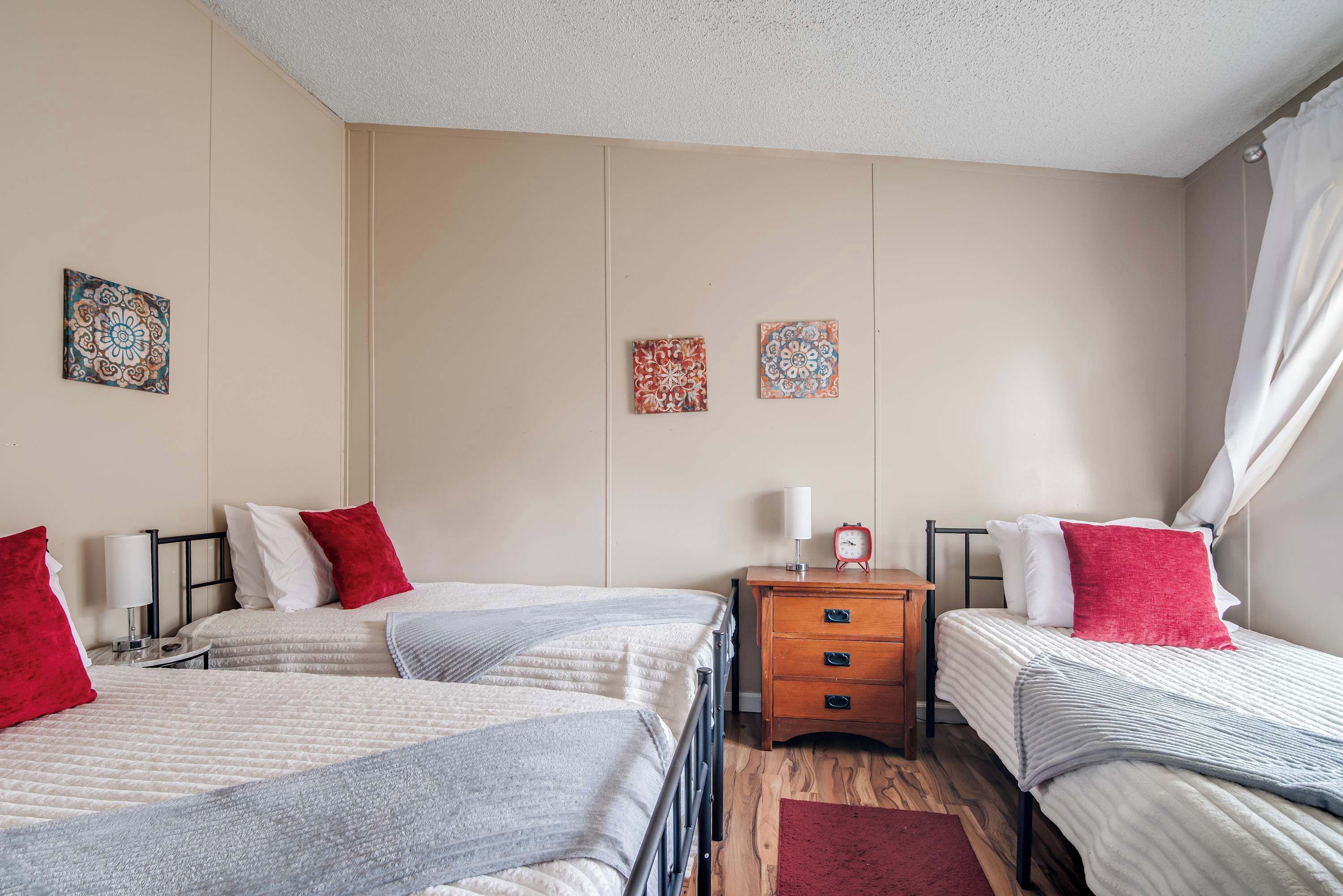 A hotel room with three beds, each with a red and white pillow, a bedside table with a lamp, and two wall-mounted decorative tiles.