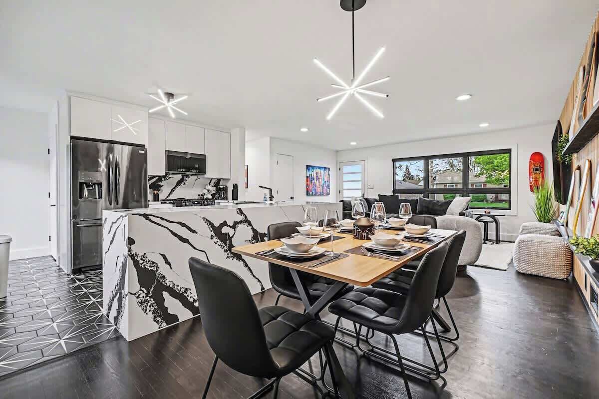 Una cocina moderna con una isla de mármol, un comedor con mesa de madera y sillas negras, y una sala de estar con sofá y televisor.