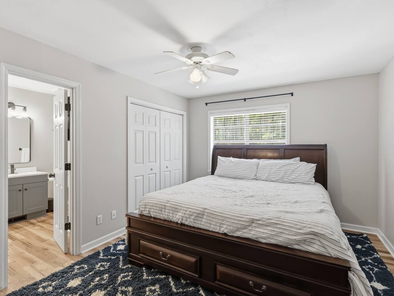 A bedroom with a bed, a ceiling fan, a window with blinds, a door, and a bathroom with a mirror and sink.