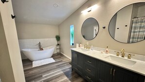A bathroom with a freestanding bathtub, a double sink vanity, dark cabinets, and a large mirror. - Remodeled Cabin in the Heart of Lakeside (Lakeside)