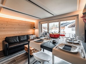 Ein modernes Interieur mit einem schwarzen Ledersofa, einem für zwei Personen gedeckten Esstisch und Blick auf eine Berglandschaft durch große Fenster.