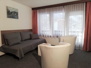Ein Hotelzimmer mit einer Couch, zwei Sesseln, einem Fenster mit durchsichtigen Vorhängen und einem Gemälde an der Wand.