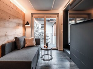 A modern interior with a wooden wall, a grey sofa with cushions, a small round table with candles, and a view through a glass door. - Kusi's Ranch Studio Studio Nr. 12 Nadelhorn by Interhome (Saas-Fee)