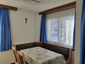 Ein Zimmer mit Tisch, einer Bank, einem Kruzifix an der Wand und einem Fenster mit Vorhängen.