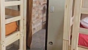 Ein schmaler Flur mit Etagenbetten an beiden Seiten, der zu einer Tür führt, von der aus man einen Raum mit Fliesenboden und Backsteinwand sieht.