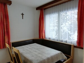 Ein Zimmer mit Tisch, Stühlen, einem Kreuz an der Wand und einem Fenster mit Vorhängen.