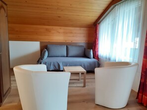 Ein gemütliches Zimmer mit Holzbalkendecke, einem grauen Sofa mit Kissen und zwei weißen Sesseln.