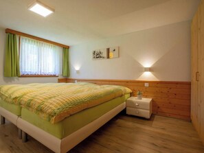 A bedroom with a bed, a bedside table, a nightstand, a window with curtains, and a wall-mounted light. - Abendstern Appartement/Fewo 5-Bettwohnung by Interhome (Saas-Almagell)