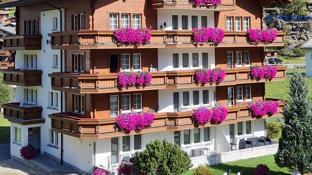 Ein mehrstöckiges Hotel mit Balkonen, die mit Blumen geschmückt sind, vor einer Bergkulisse.
