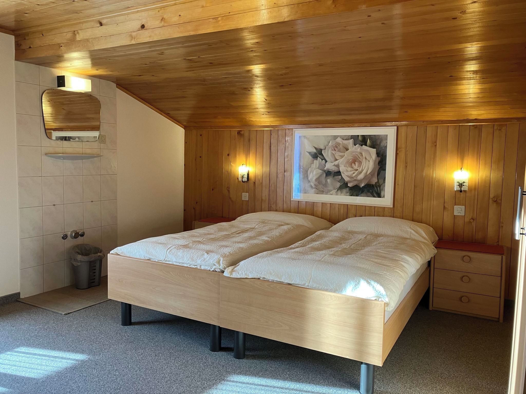 Ein Schlafzimmer mit Holzbalkendecke, einem Bett mit weißer Bettwäsche, zwei Nachttischen und einer an der Wand befestigten Leuchte.