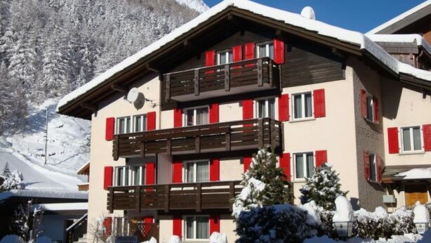 Un edificio de varios pisos con balcones, un paisaje nevado y montañas al fondo.