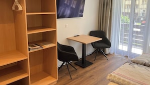 Una habitación moderna con una estantería de madera, una mesita con una silla, un televisor montado en la pared y un ventanal con cortinas.