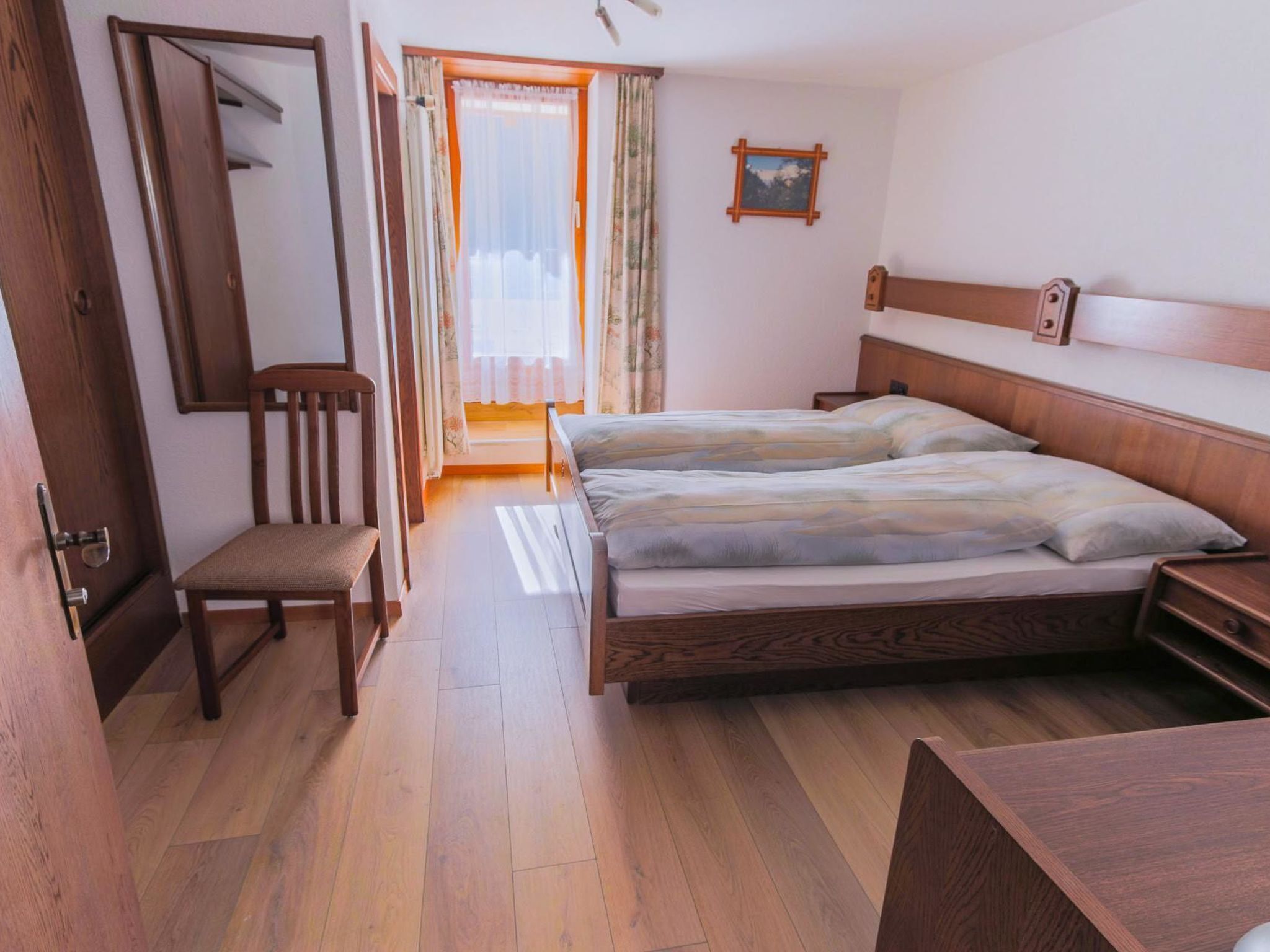 A bedroom with a wooden bed, a chair, a bedside table, and a window with curtains.