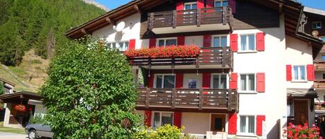 Un immeuble à plusieurs étages doté de balcons, avec un arbre au premier plan et une chaîne de montagnes en arrière-plan.