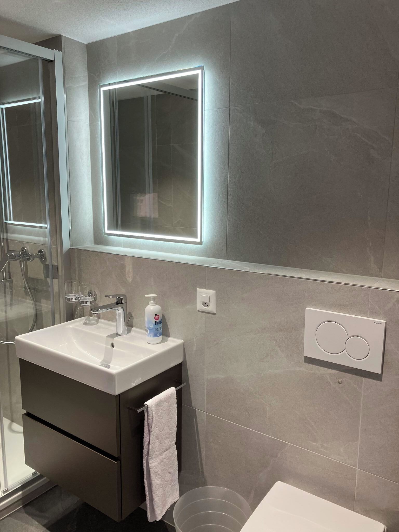 Ein modernes Badezimmer mit einer Duschkabine aus Glas, einem weißen Waschbecken und einem Spiegel mit integrierter Beleuchtung.