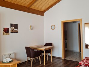 Ein Essbereich mit Holztisch und -stühlen, einer Wanduhr und einer durch einen offenen Durchgang einsehbaren Küche.