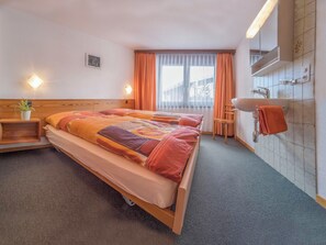 A hotel room with a bed, a bedside table with a plant, a bathroom with a sink and mirror, and a window with curtains. - Bergrose Appartement/Fewo 4-Bettwohnung Nr. 3 by Interhome (Saas-Fee)