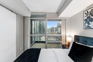 A hotel room with a bed, a nightstand, a lamp, a large window with a city view, and a door leading to a balcony. - Altitude Residences (Toronto)