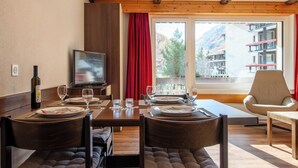A dining area with a wooden table, chairs, a television, and a window with a view of buildings and trees. - Haus Azur Appartement/Fewo Wohnung Nr. 1 by Interhome (Saas-Almagell)