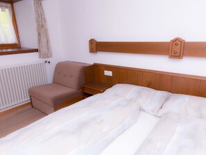 Quarto com cama, mesa de cabeceira, cadeira, janela com cortinas e radiador.