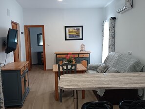Ein Wohnzimmer mit einer Couch, einem Esstisch mit Stühlen, einem an der Wand montierten Fernseher und einer kleinen Kommode.