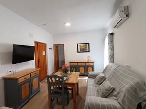 Un saló amb un sofà, una televisió penjada a la paret, una taula de dinar amb caires i una zona de cuina amb armaris.