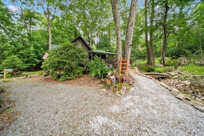 A rustic cabin surrounded by dense forest, a gravel driveway, and a wooden bridge. - Creekside Escape with Hot Tub & Outdoor Fireplace (Banner Elk)